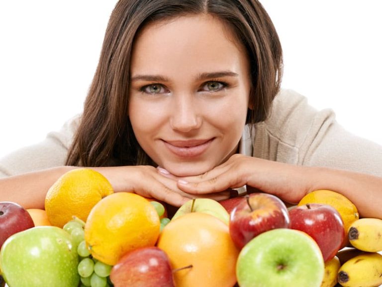 Smiling woman with glowing skin and an array of healthy fruit to illustrate best foods for your skin